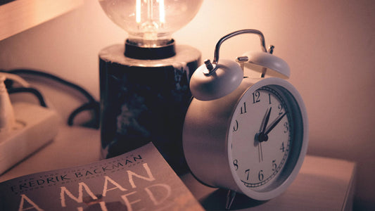 An alarm clock beside a book and a lamp placed on a bedside table.