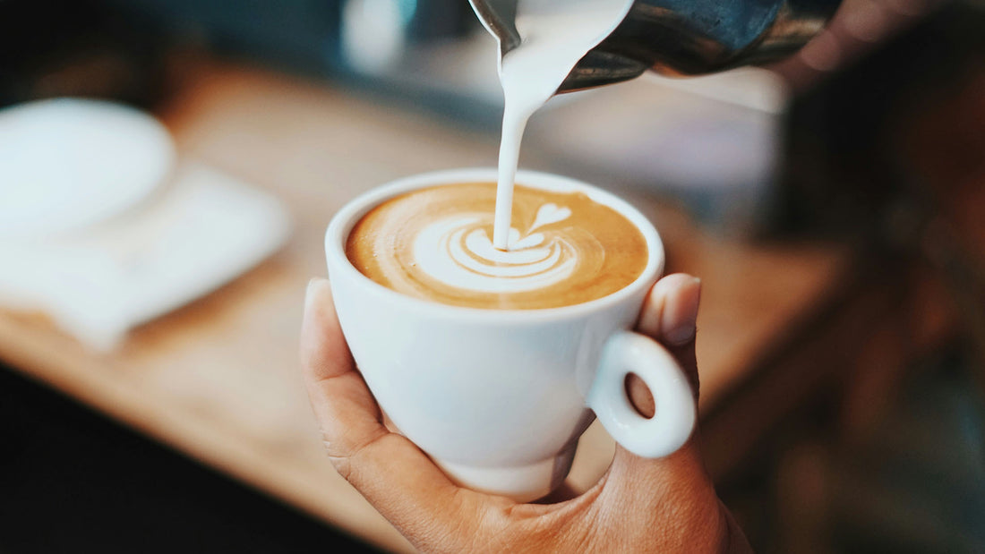 Coffee cup in a hand with milk being poured into the almost full cup. Milk forms a pattern on the top of the coffee, forming a small heart with three ripples around it.