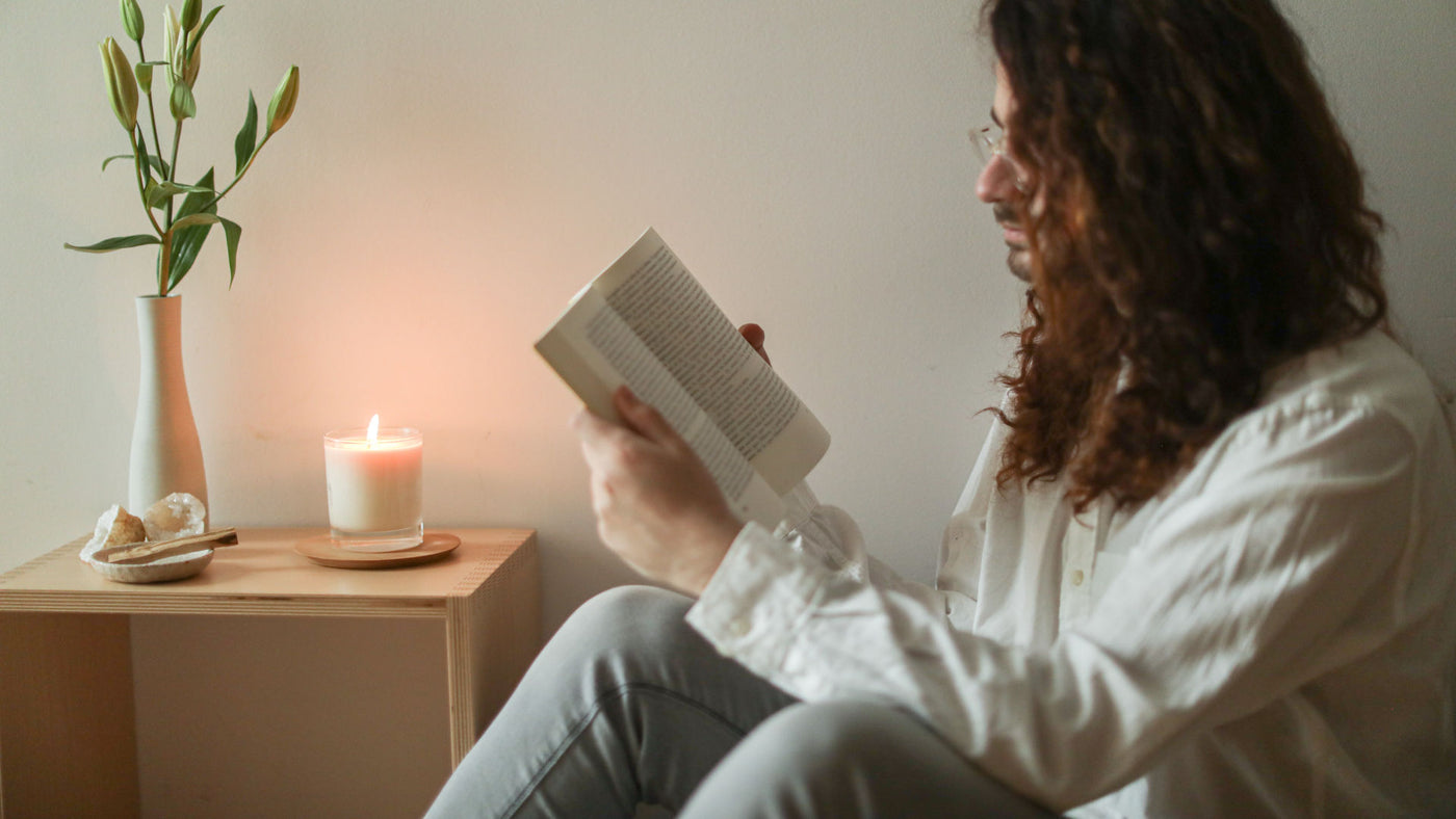 A long-haired man sitting on the floor reading a book with a lighted scented candle on the side table.