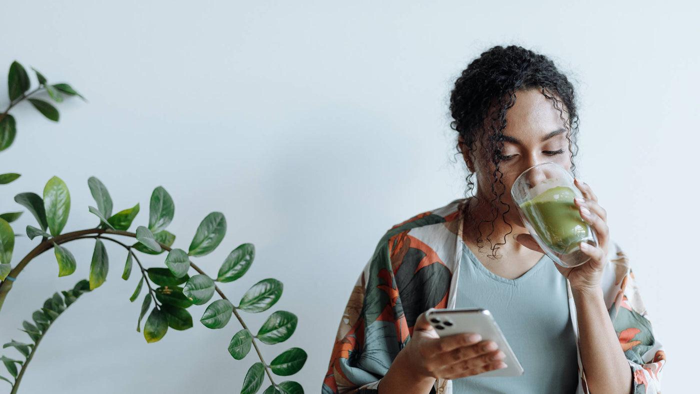 A woman of color drinks a green matcha latte while checking her smartphone.