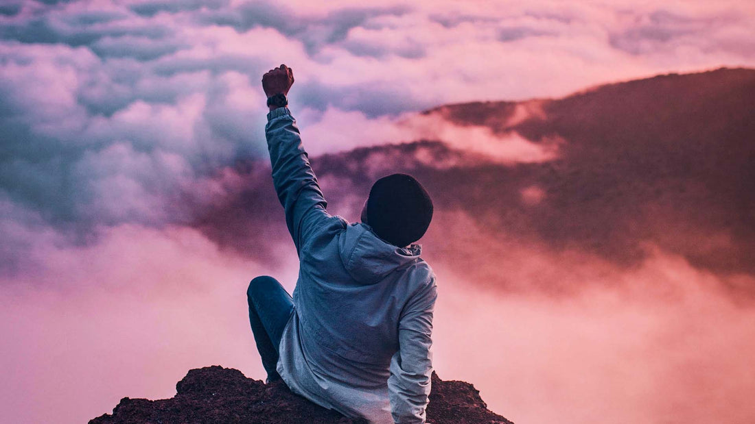 Man sitting on mountain cliff facing white clouds rising one hand at sunset