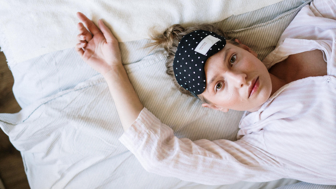 A woman lying on a white bed and wearing a sleep mask on her forehead.