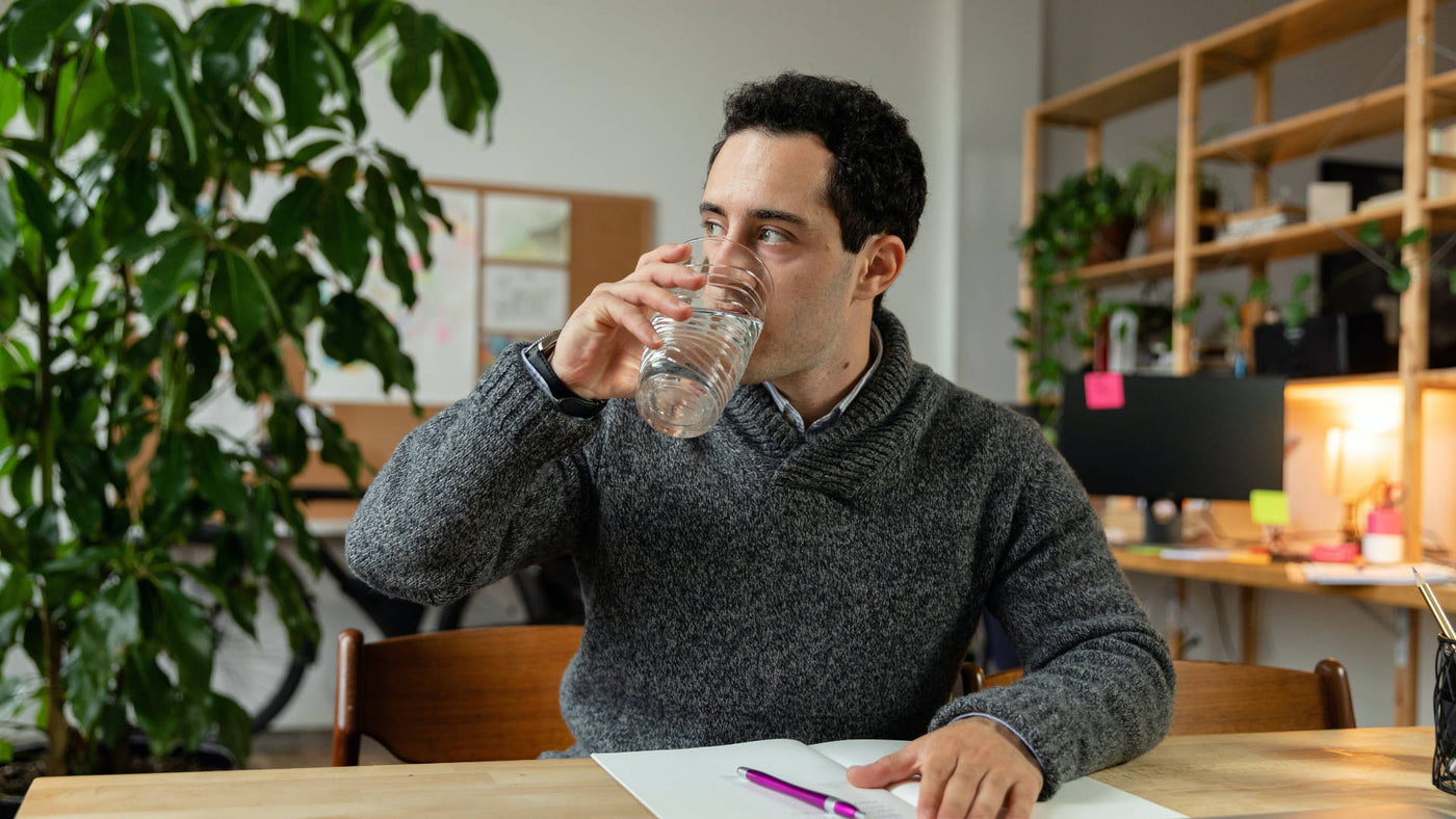 A man drinking a glass of water looking at his side while sitting on his office desk.