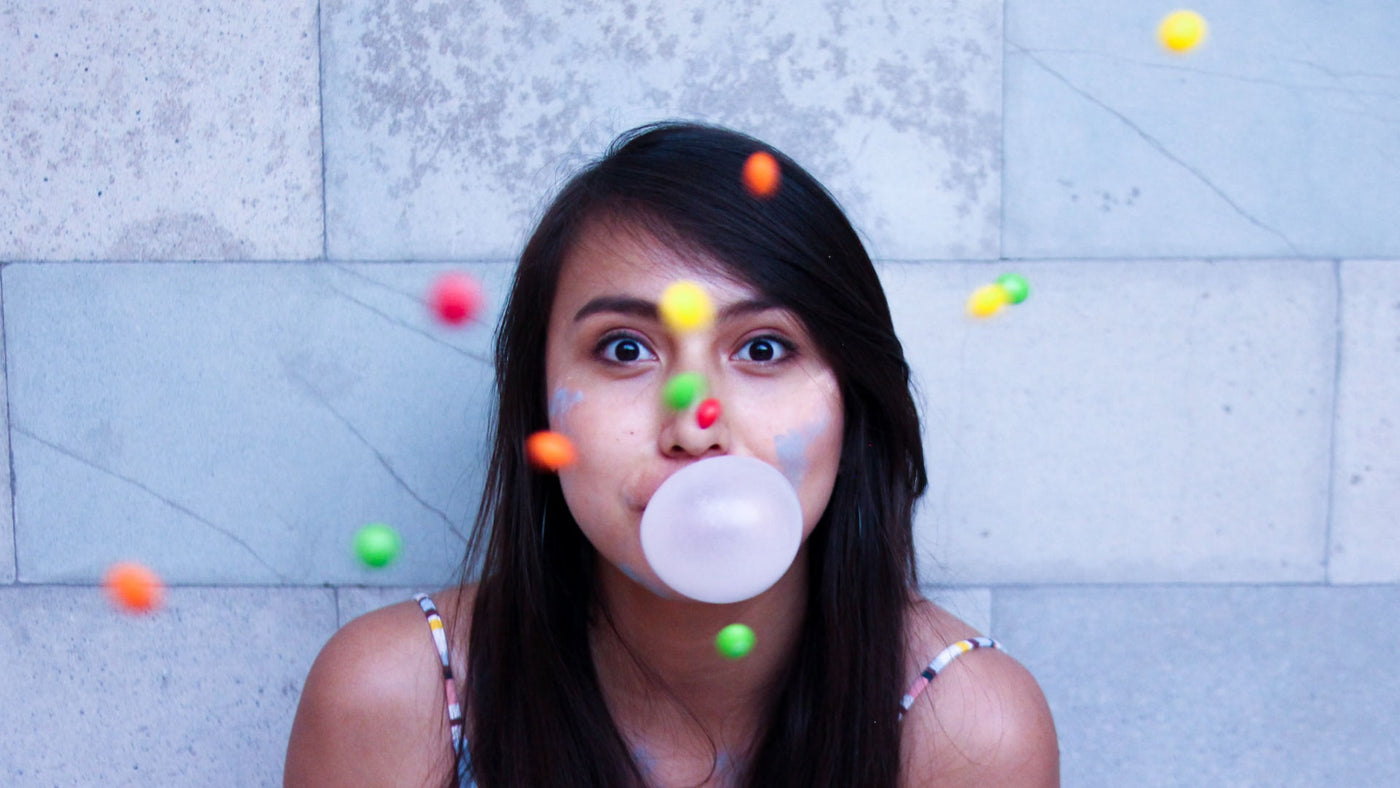 A young, playful woman making a gum bubble with colorful candies dropping like confetti in front of her.