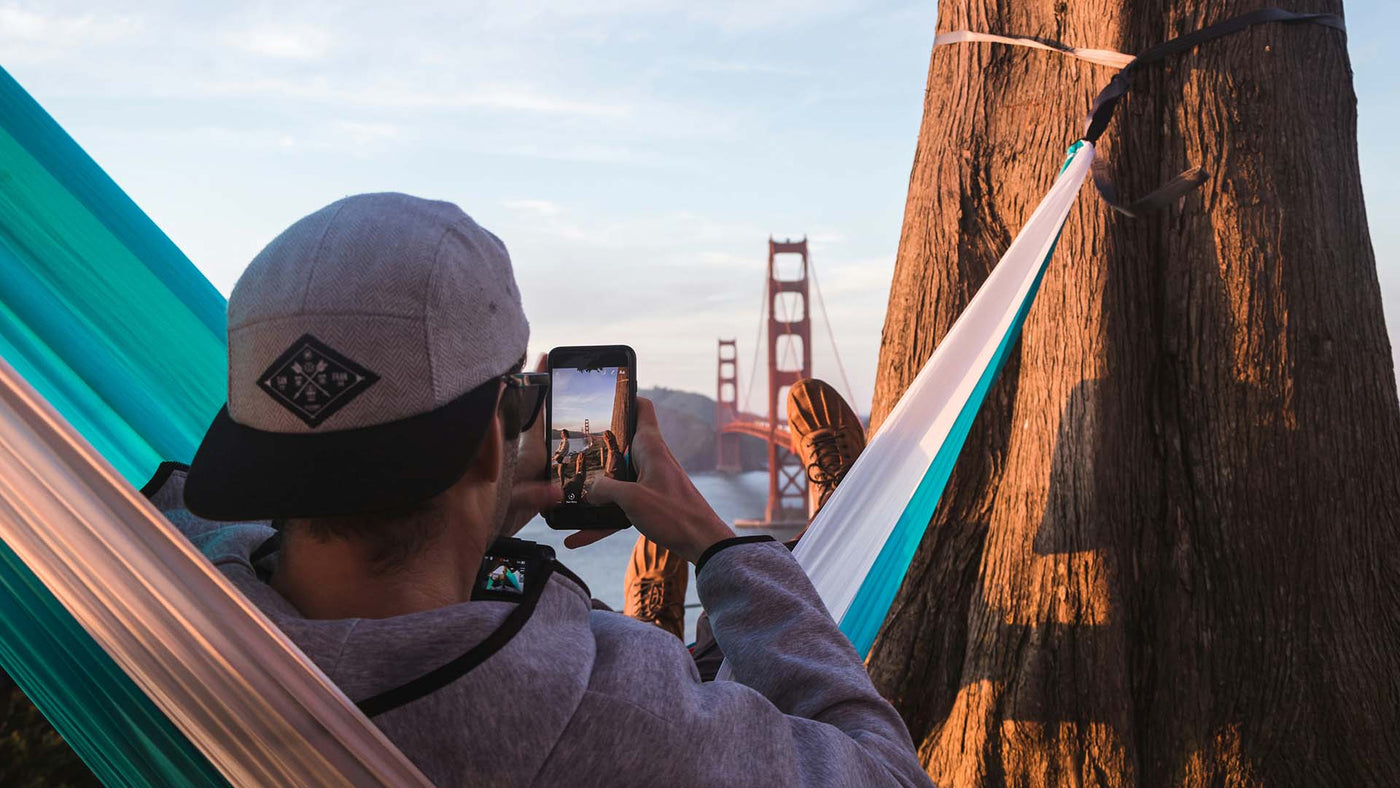 shallow focus photography of man on hammock while taking photo of golden gate bridge of california to his phone