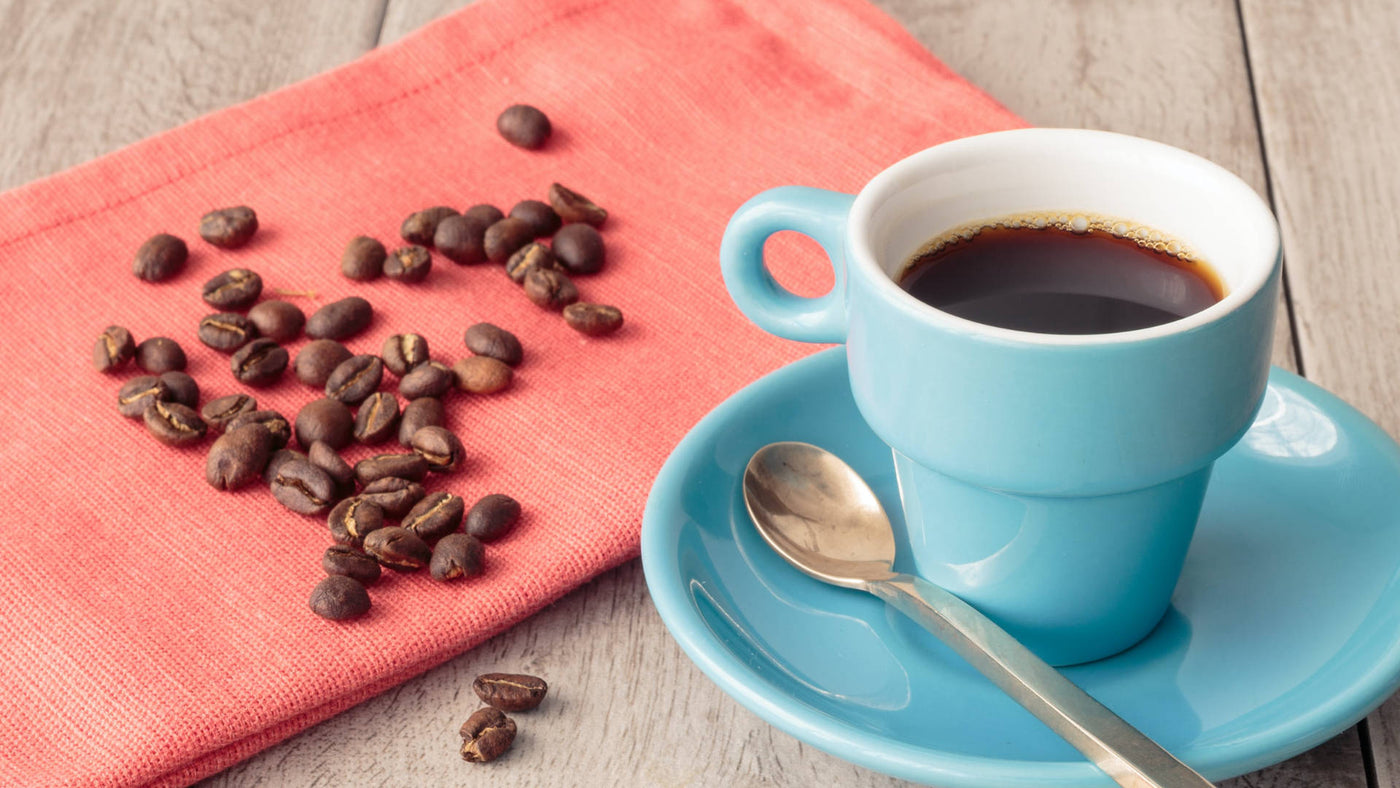 A blue cup with saucer filled with black coffee and coffee beans scattered on a pale red kitchen towel. 