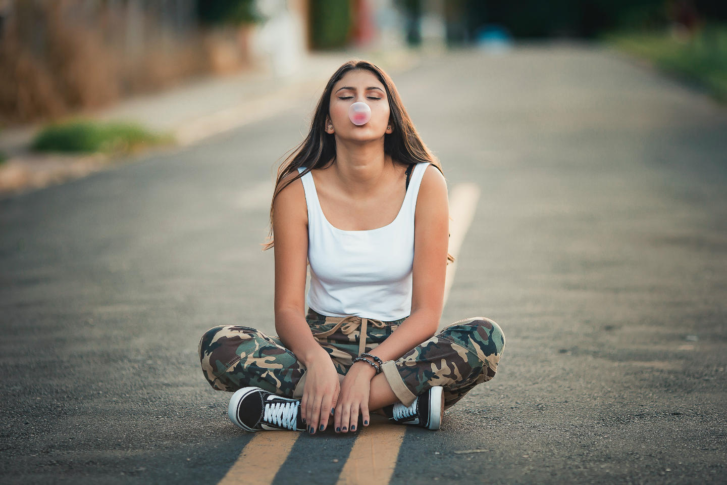 A young woman sitting in the middle of the road with her legs crossed while blowing a bubble gum.