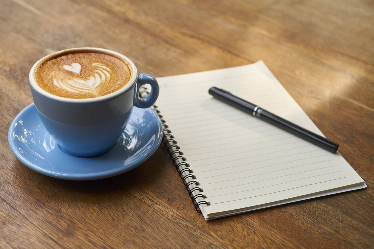 A coffee heart-shaped latte art in a blue cup beside a blank notebook and pen on a wooden surface.