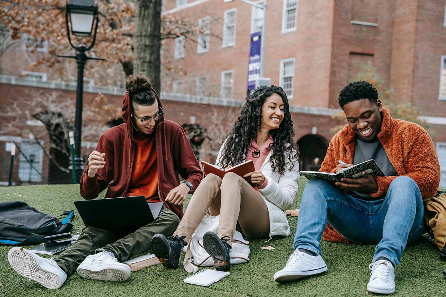 three college students sitting on the grass with laptop and books