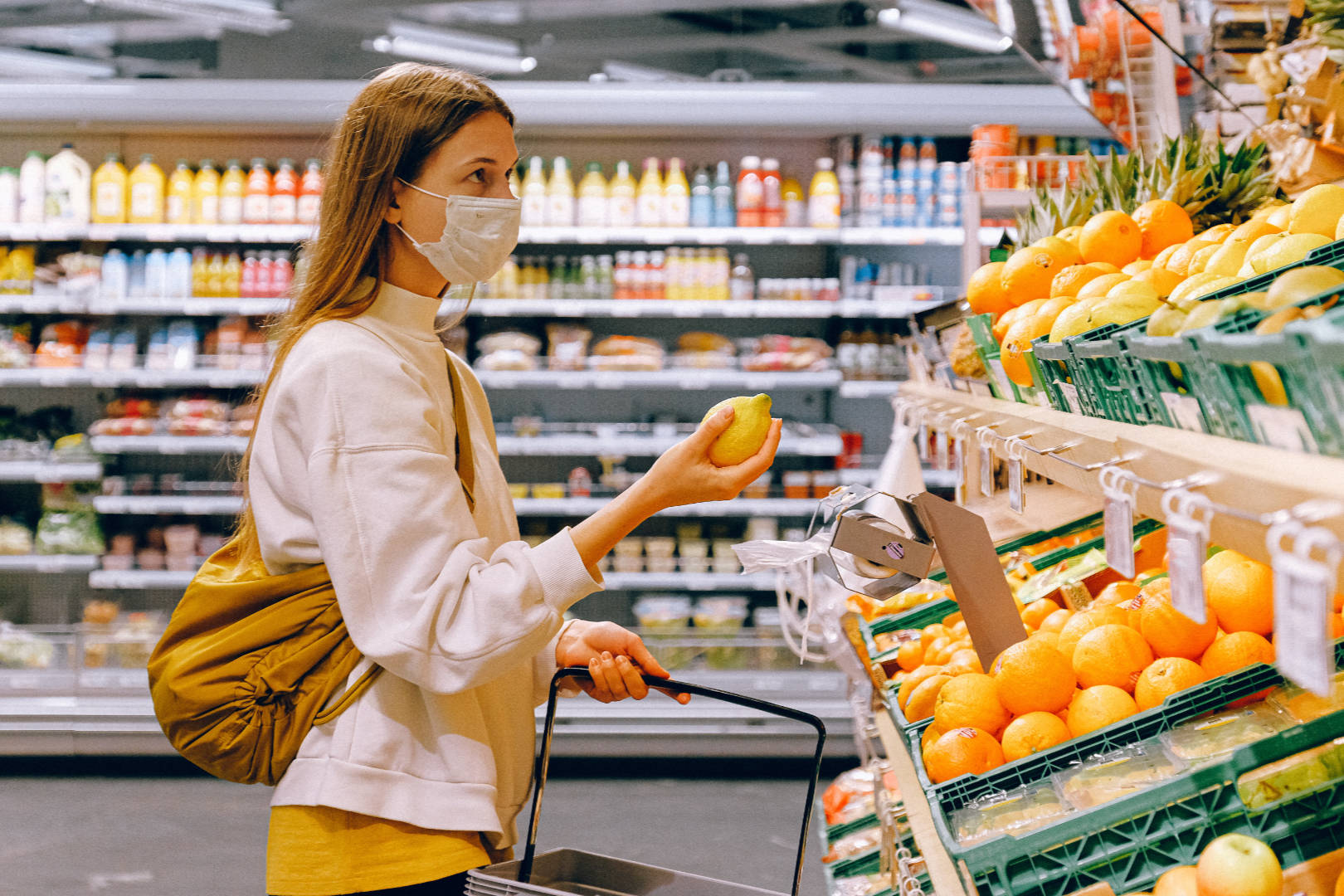 A young woman wearing a face mask holding a shopping cart while selecting fruits on a grocery.