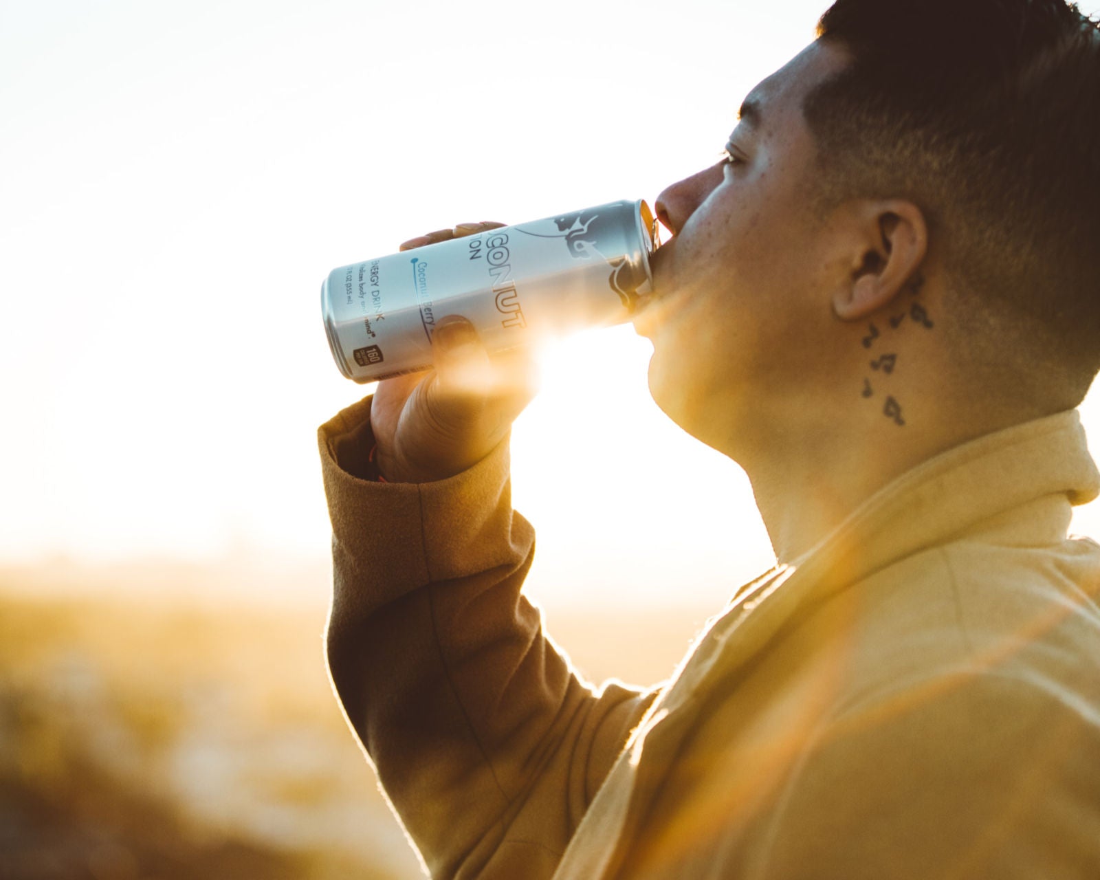 A man wearing a yellow coat while drinking an energy drink from can with sunlight peeking in the background.