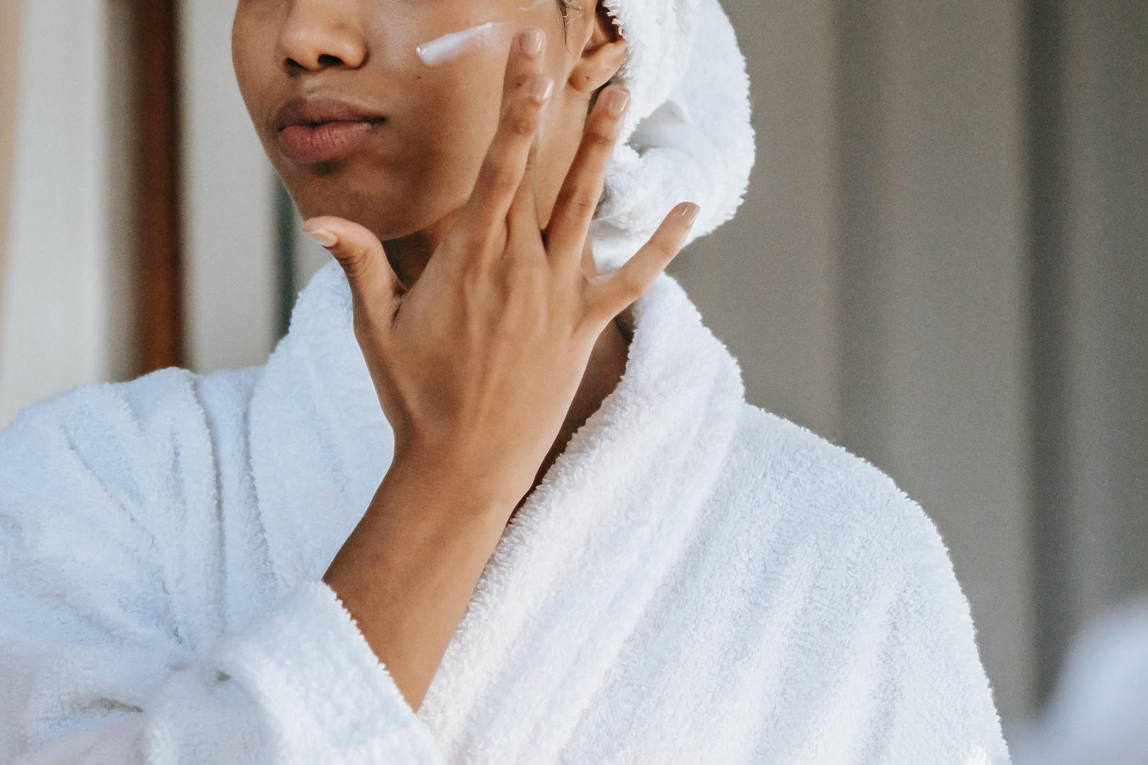 A woman of color applying face moisturizer on her left cheek.
