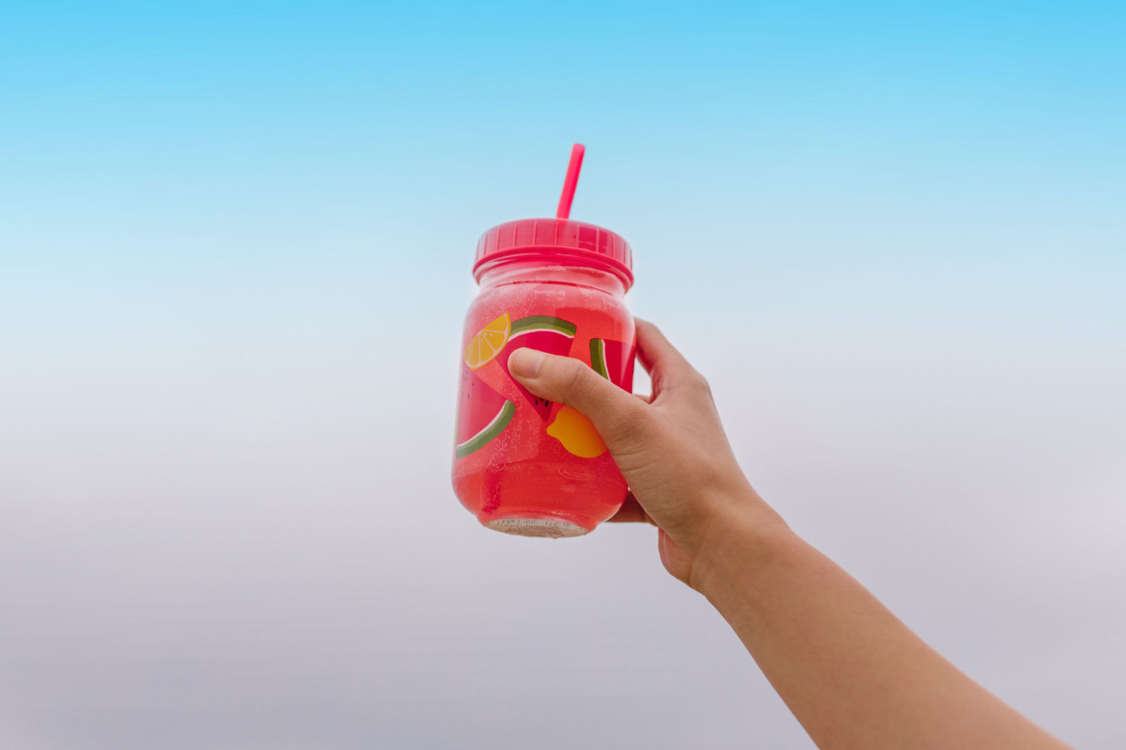 A person holding a glass of watermelon juice up in the air while showing the bright sky on the background.