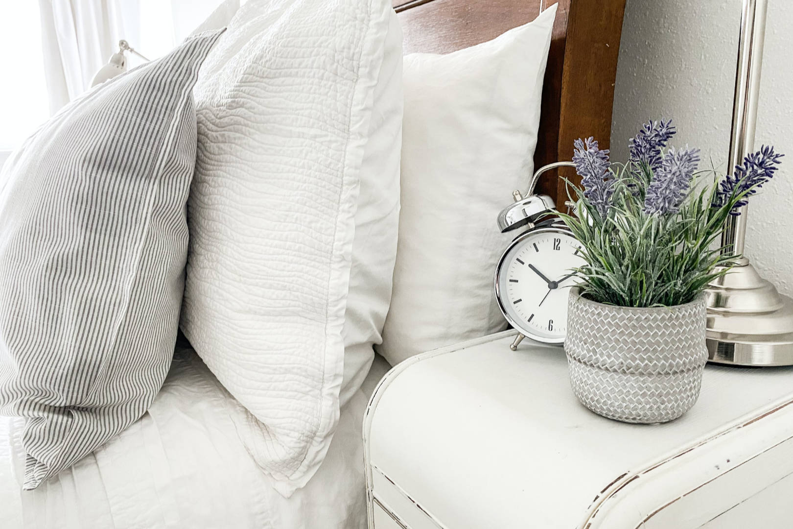 A bed with several pillows and a bedside table with alarm clock and plant placed on top.