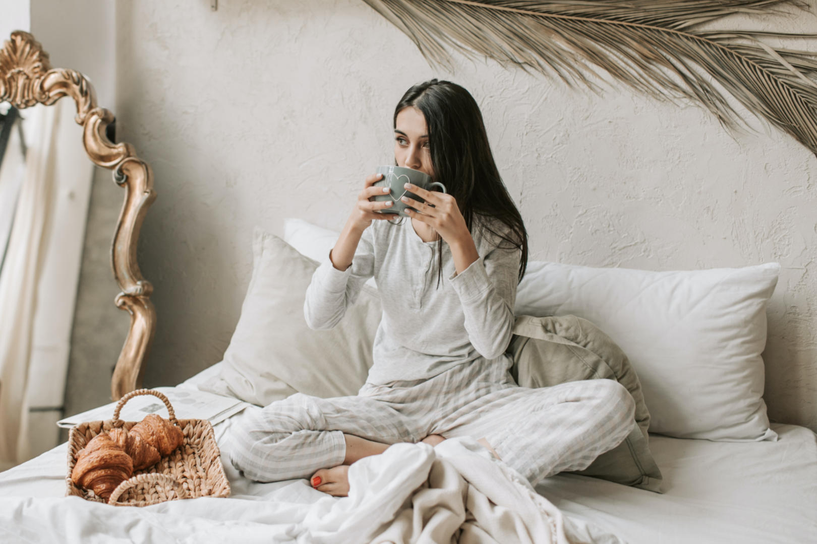 A young woman drinks from a cup while sitting on her bed.