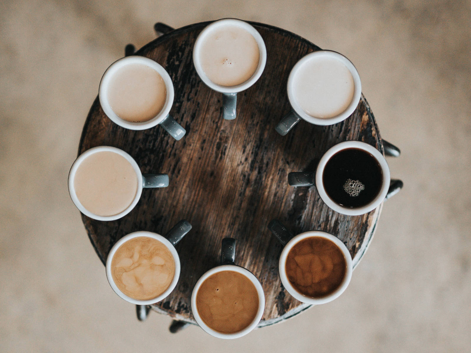 A flat lay shot of eight different types of coffee drinks ranging from pure black, latte, cappuccino to flat white in mugs on round table.