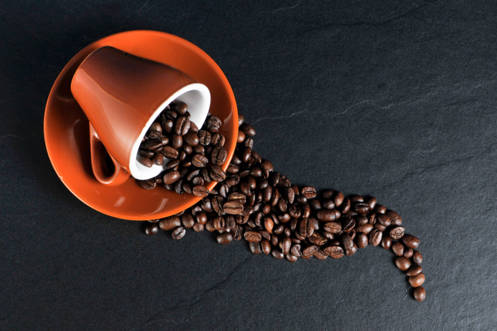 Coffee beans spilled on a black surface from an orange ceramic mug.
