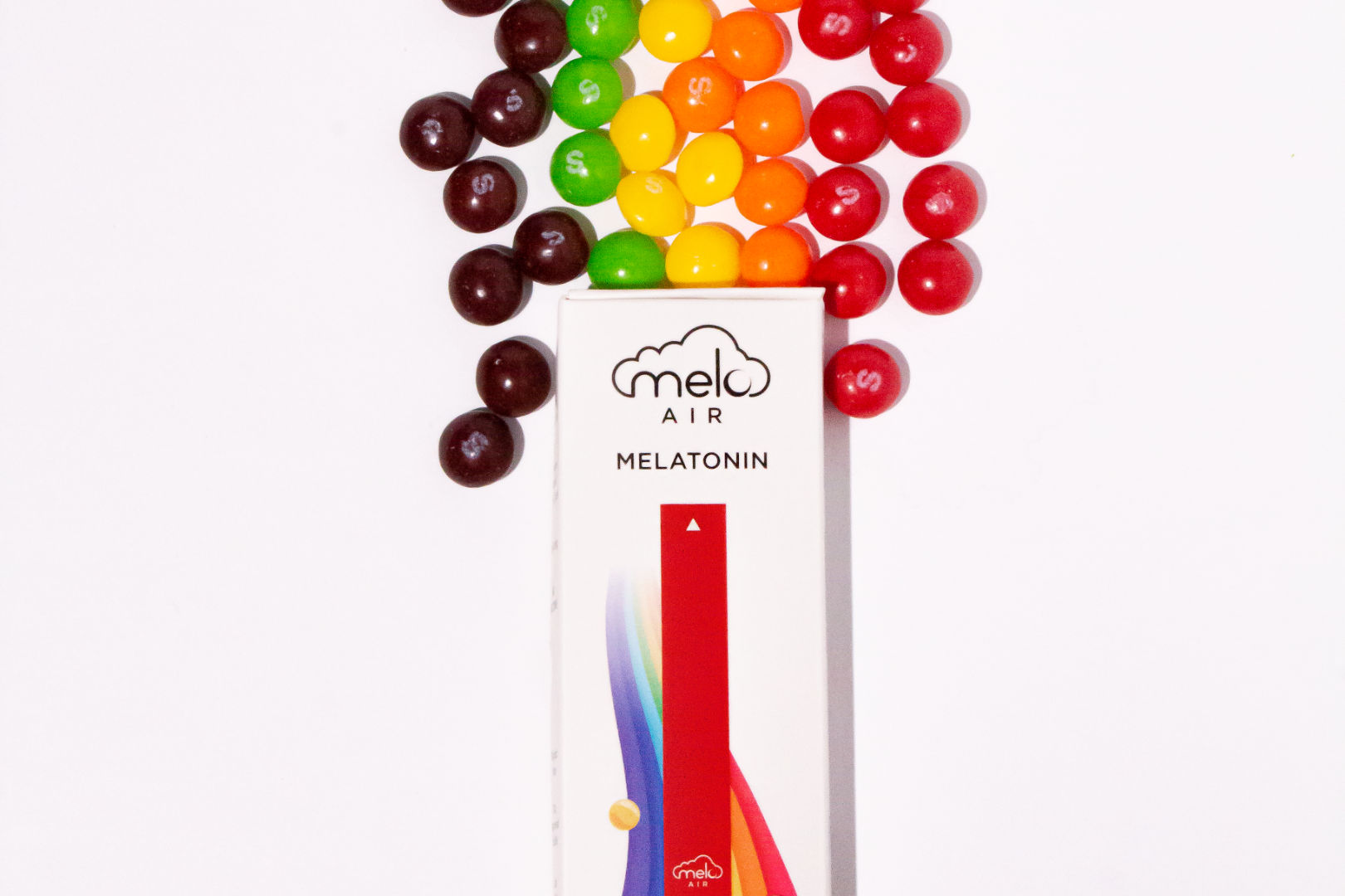 A box of Melo Air Melatonin Diffuser in Rainbow Road flavor with chocolate candies lined up in rainbow colors behind it.