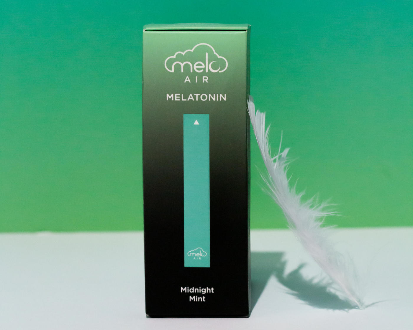 A box of Melo Air Melatonin Diffuser in Midnight Mint flavor and a white feather leaning on its right side in a cool green background