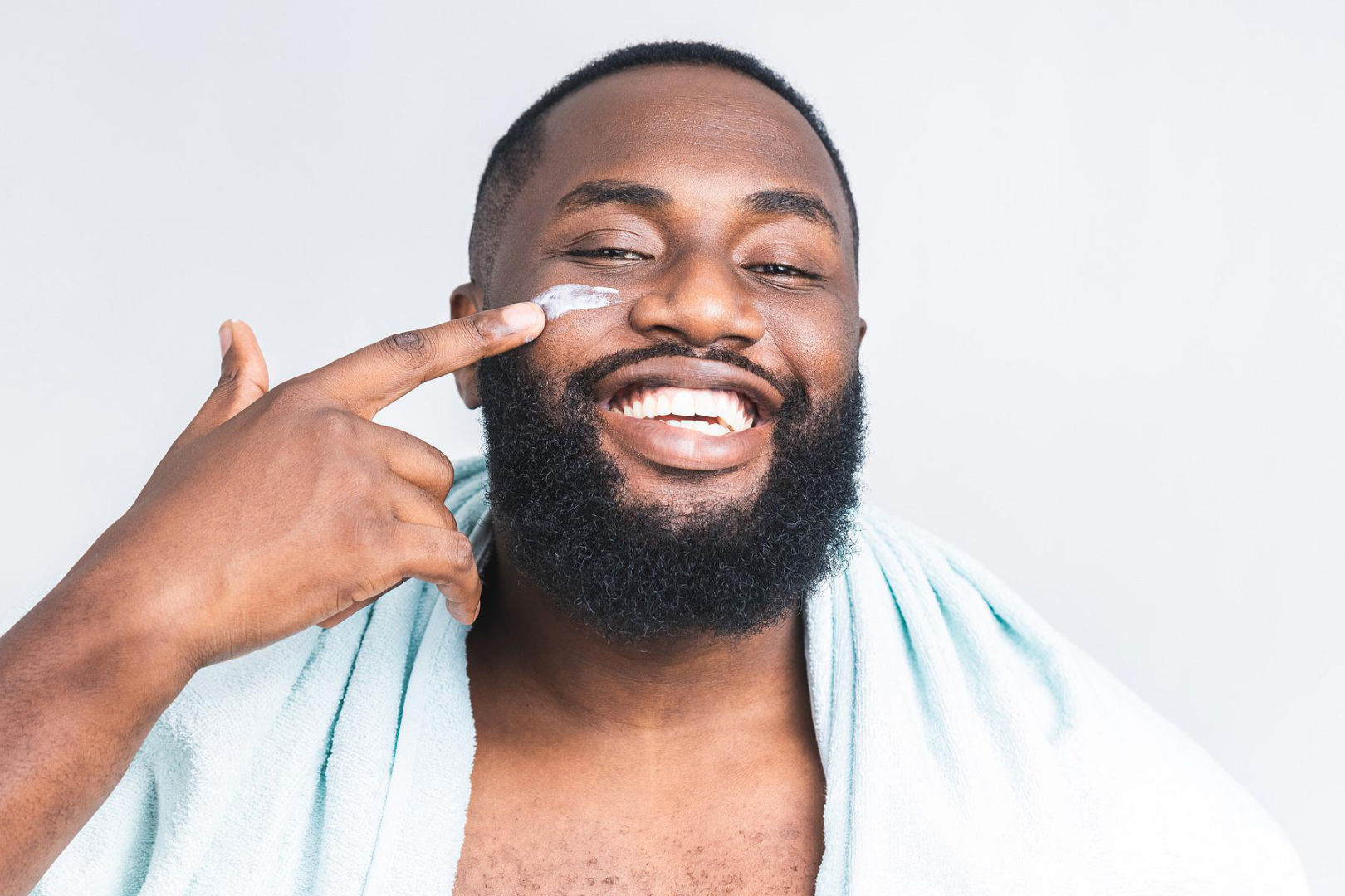 smiling man applying a skin care cream on his right cheek using his right index finger