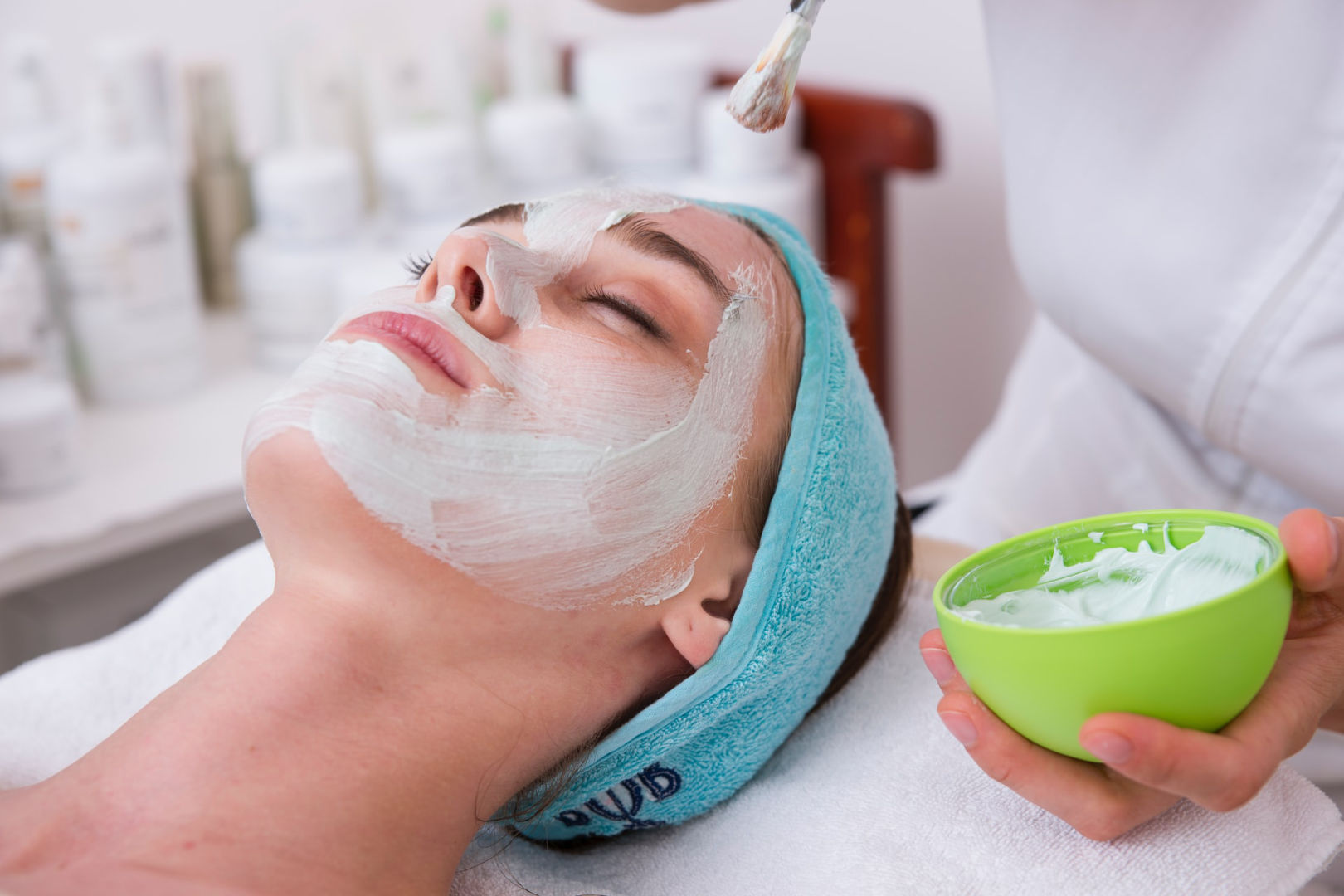 A woman sitting on a facial chair while an esthetician is applying a cream all over her face.