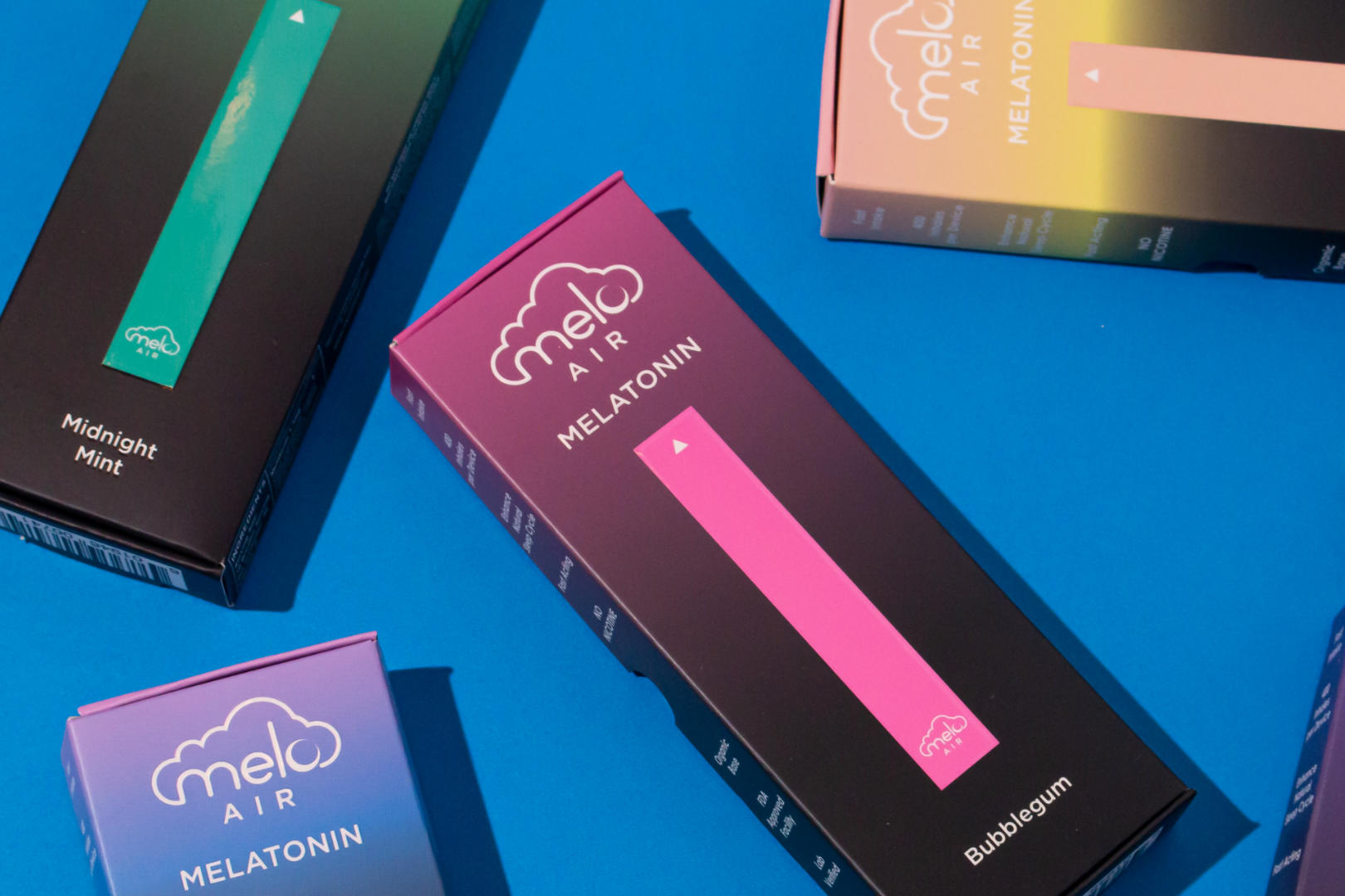 Several boxes of MELO Air Melatonin Diffusers in bubblegum, midnight mint, and peach cream flavors scattered in a blue surface.