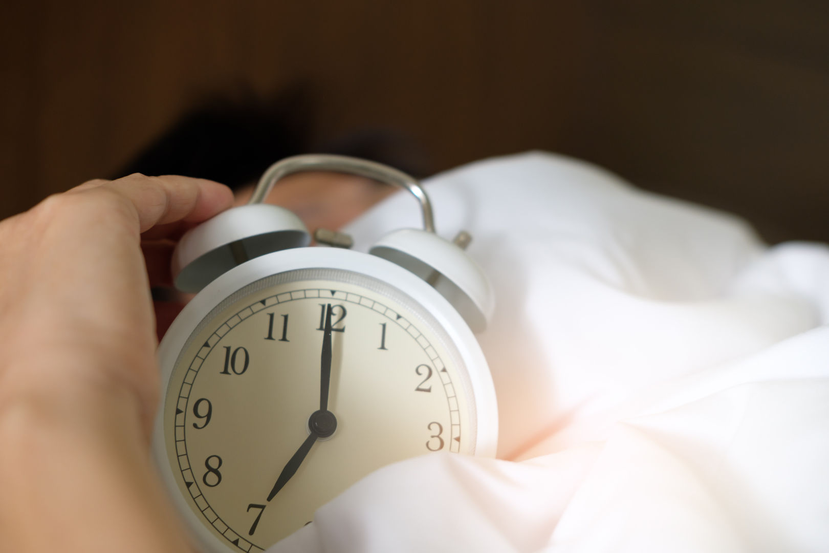 A person holding an alarm clock while another person is sleeping behind.