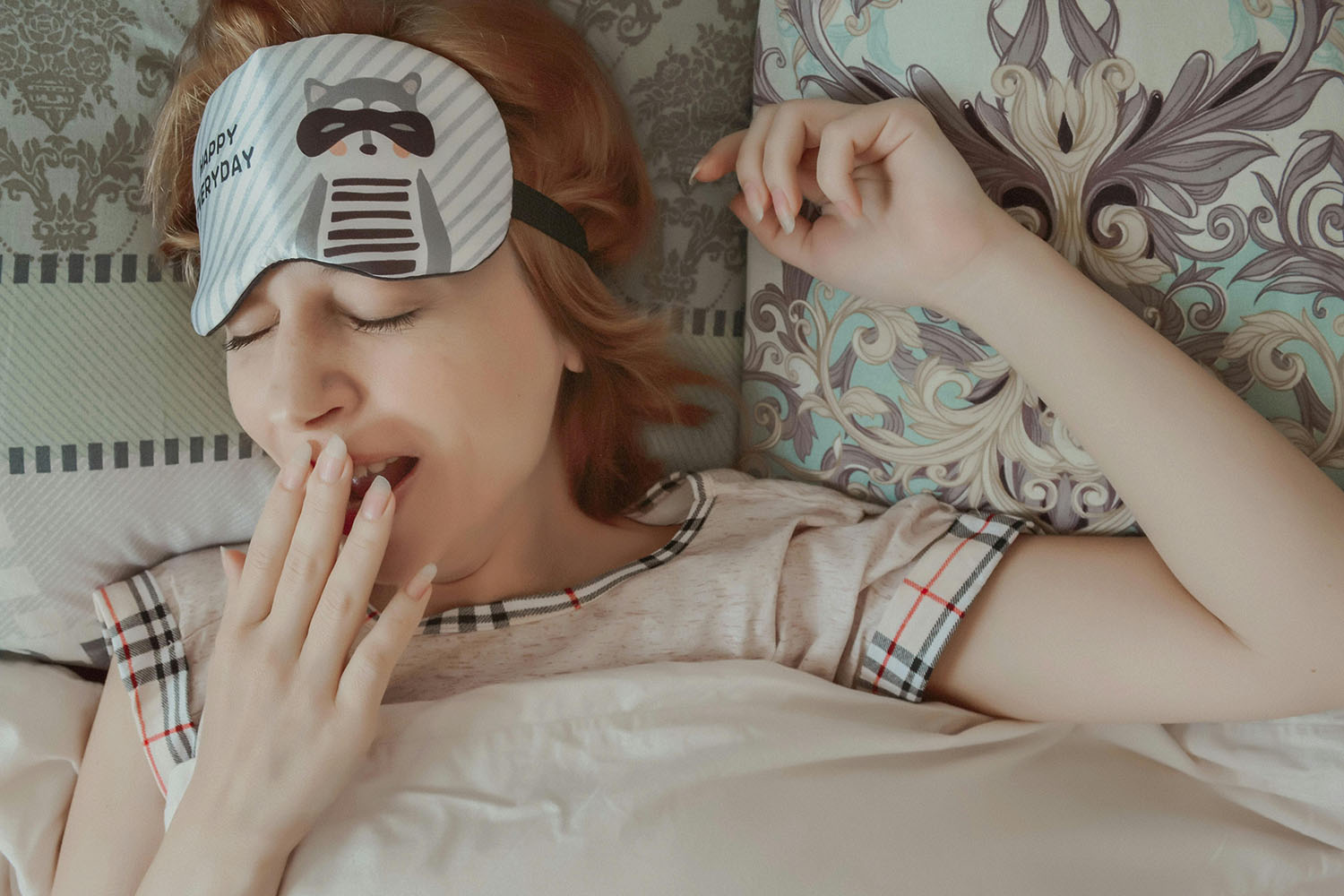 A woman in bed yawning with an eye mask resting on her forehead