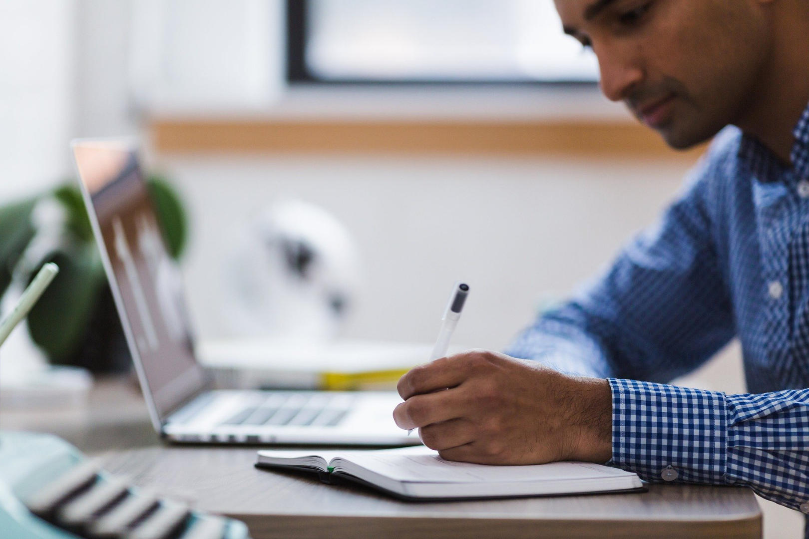 A man writing on a journal with a laptop on table.
