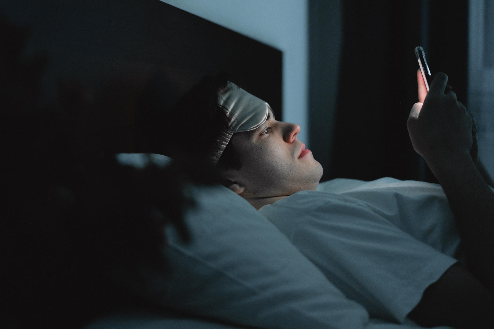 A man with an eye mask looking at his smartphone while lying in bed at night.