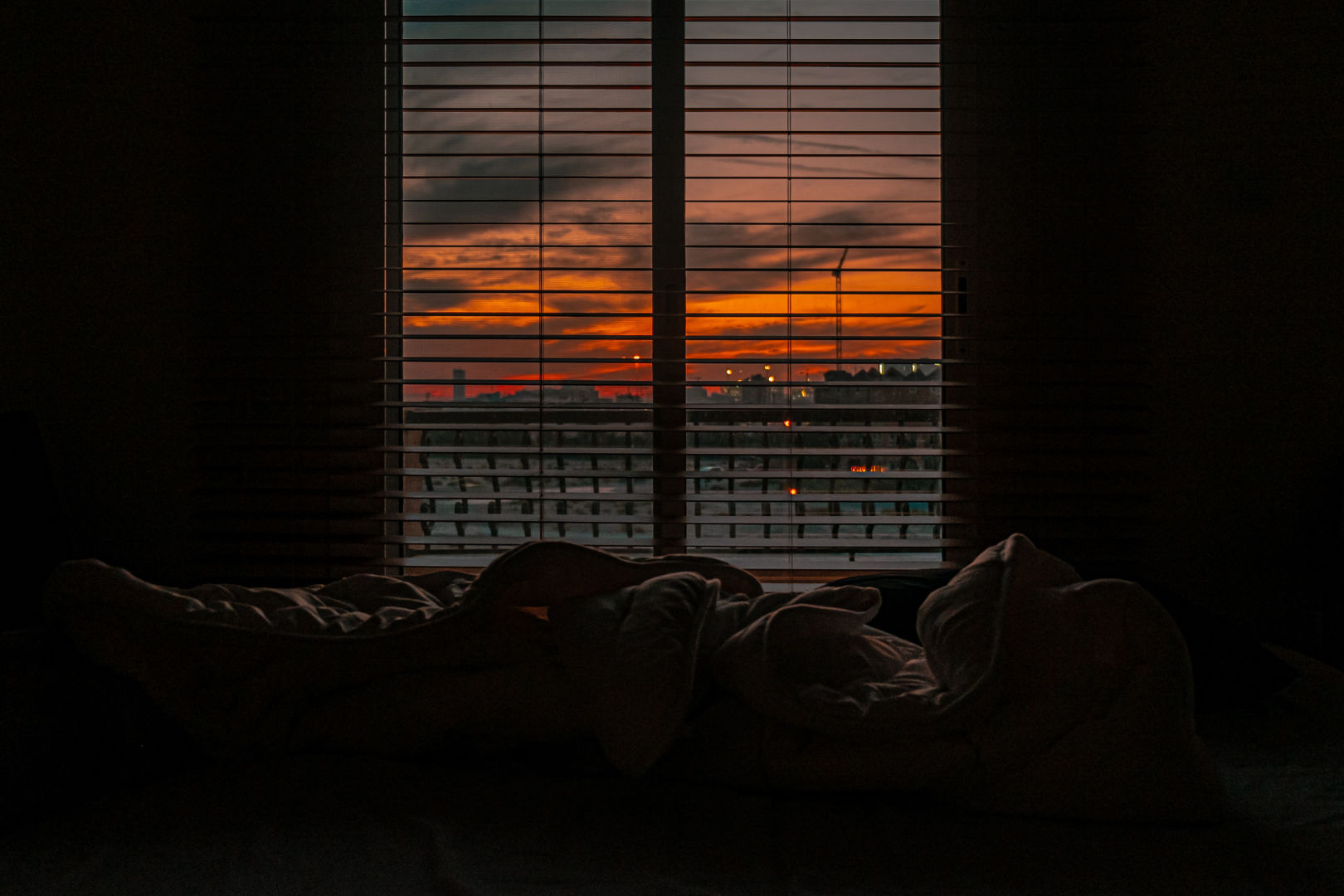 A dark bedroom with bedsheets scattered on the bed and an open window blinds showing the burnt orange sunset outside.