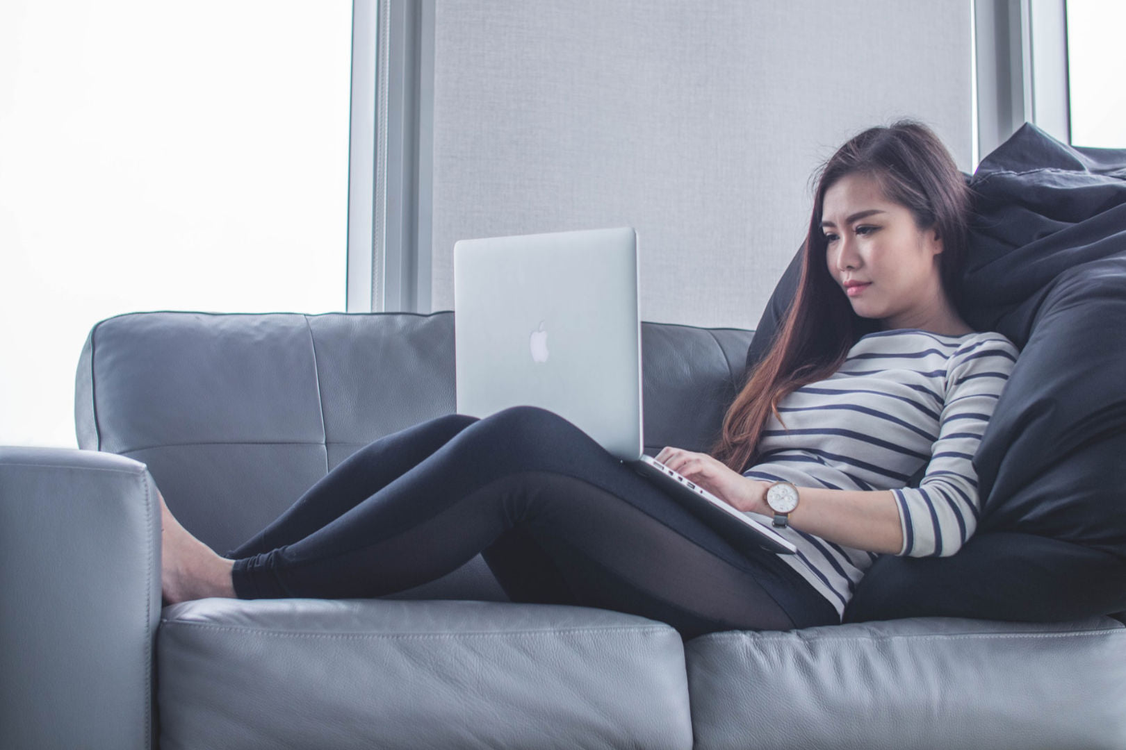 A woman sitting on a blue grey sofa while using a MacBook.