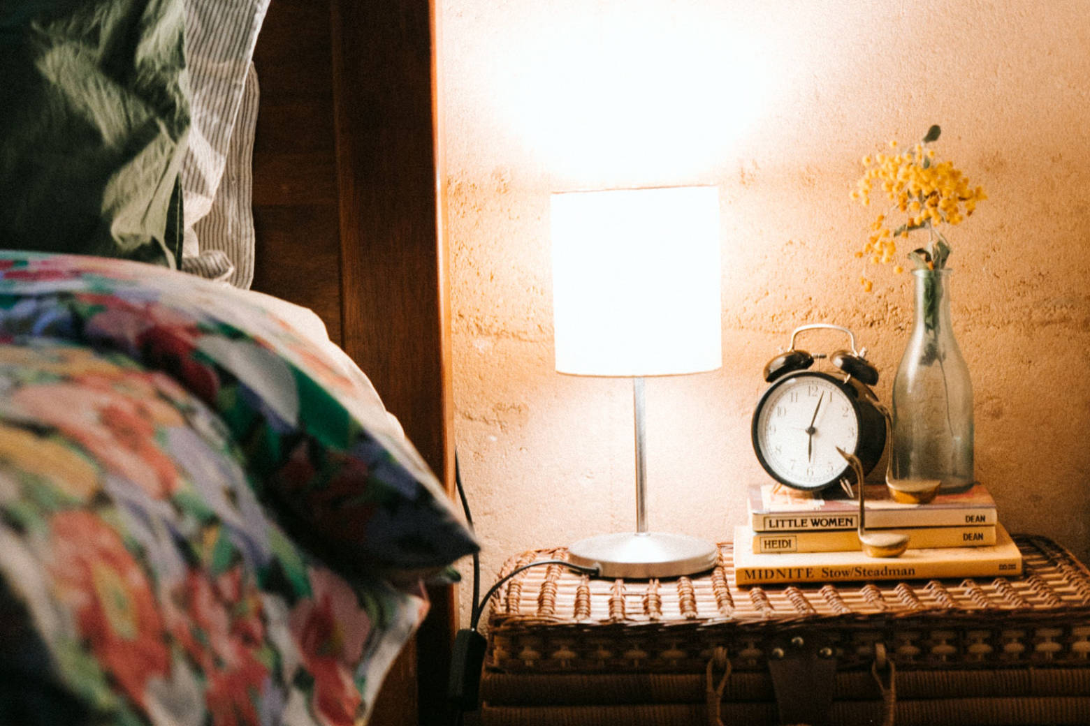 A rustic luggage used as a bedside table with alarm clock and lamp on top beside a bed.