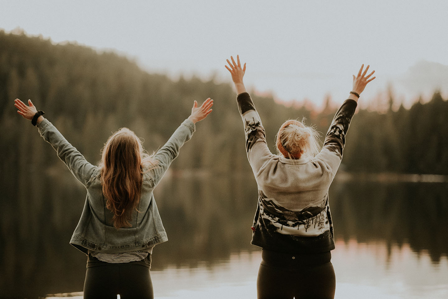 two women standing by a lake, joyfully raising their arms in celebration