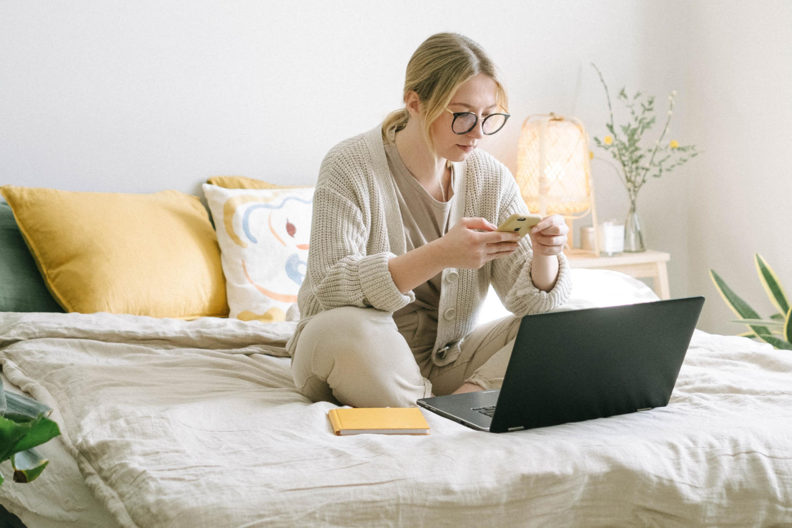 A woman sitting on her bed while looking at her smartphone and with a laptop open